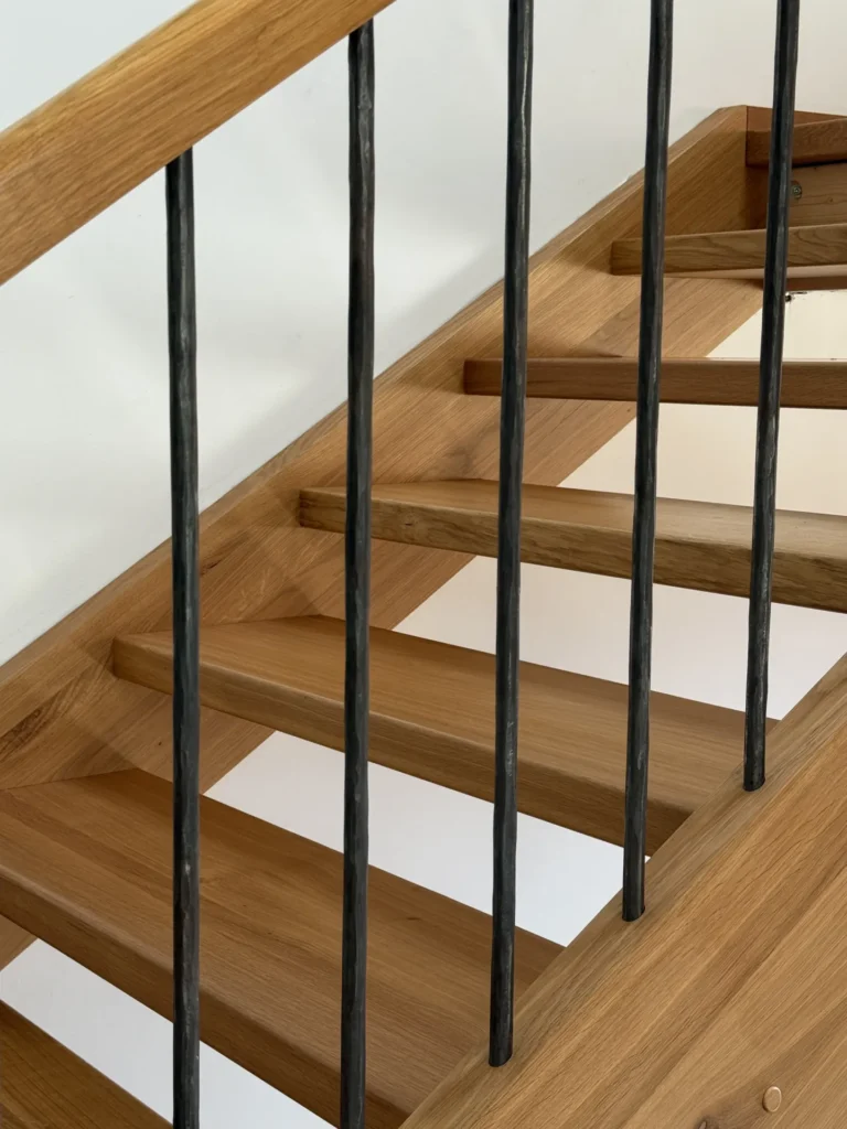 Nahaufnahme einer modernen Holztreppe mit vertikalen Geländern aus schwarzem Metall und einer weißen Wand im Hintergrund. Die Stufen und das Geländer bestehen aus hellem Holz.