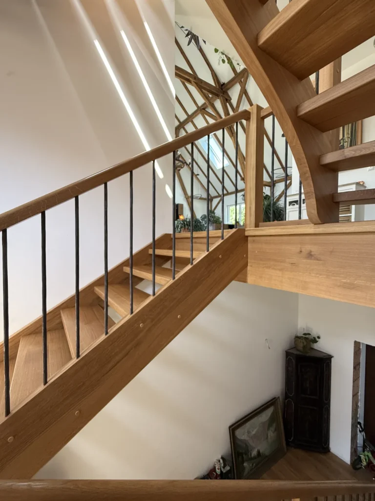 Eine moderne Holztreppe mit schwarzen Metallgeländern, Sonnenlicht, das durch schräge Fenster hereinströmt, und freiliegende Holzbalken an der Decke. Unten stehen ein Schrank und gerahmte Kunstwerke, die an der weißen Wand lehnen.