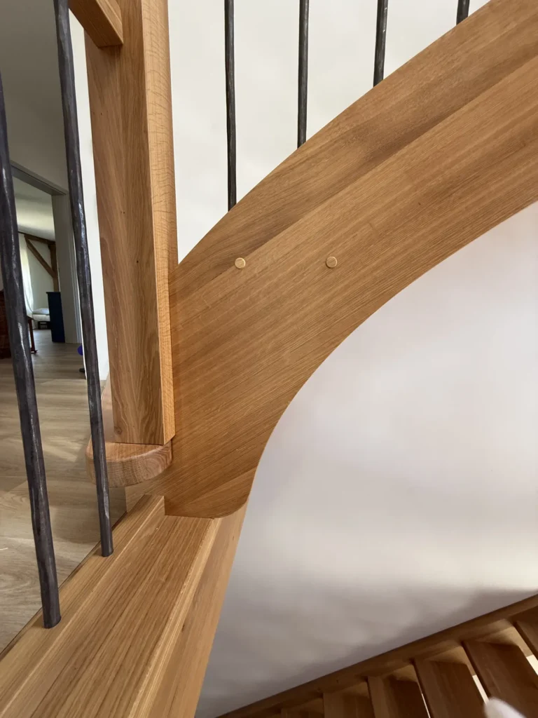 Nahaufnahme einer Holztreppe mit Metallbalustern, die die detailreiche Tischlerarbeit und die glatten, geschwungenen Holzformen vor einer weißen Wand in einem modernen Interieur zeigt.
