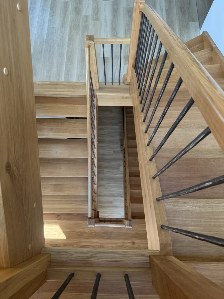 Blick hinunter auf eine Holztreppe mit schwarzen Metallgeländern, die mehrere Treppenläufe und Podeste zeigt, sowie einen hellen Holzboden am Fuß der Treppe. Das Bild betont geometrische Linien und Tiefe.