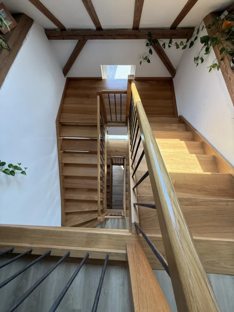 Blick hinunter auf eine moderne Holztreppe mit Metallgeländer, umgeben von weißen Wänden und Pflanzen. Sonnenlicht strömt durch ein Fenster oben herein und beleuchtet die Stufen und die Holzdetails.