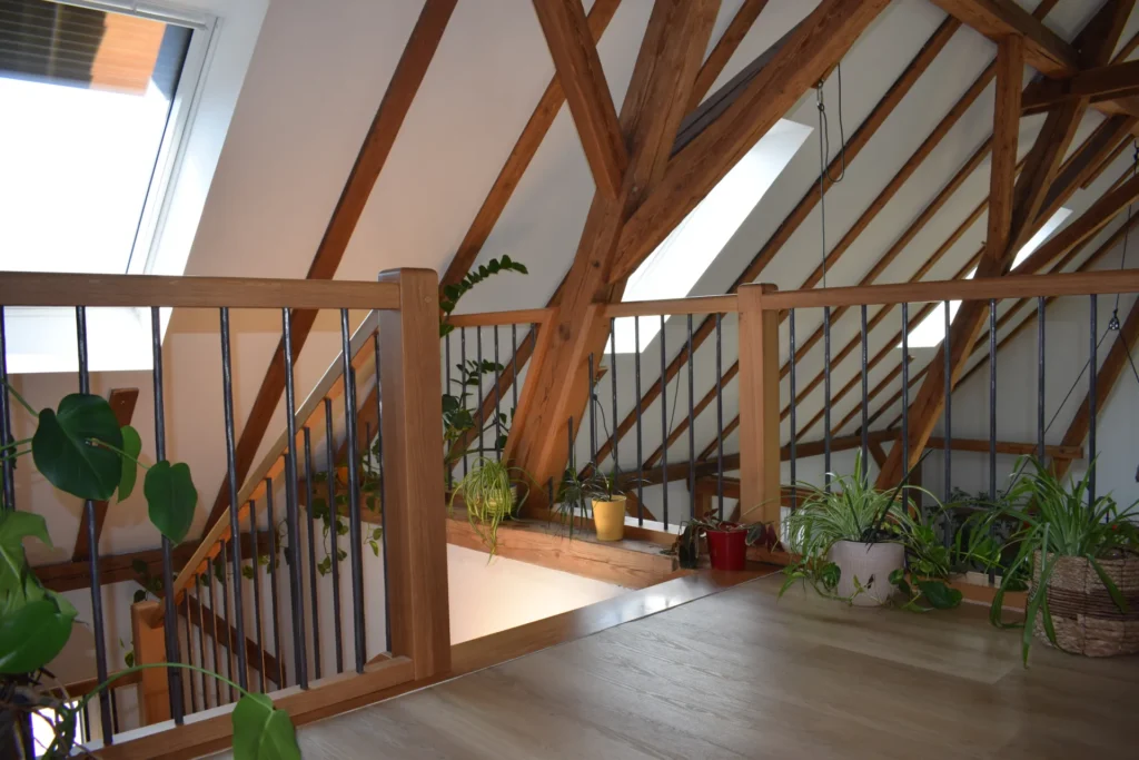 Ein Loft mit Holzbalken, Metallgeländern und Topfpflanzen befindet sich unter einer schrägen Decke mit großen Fenstern, durch die Tageslicht hereinströmt.