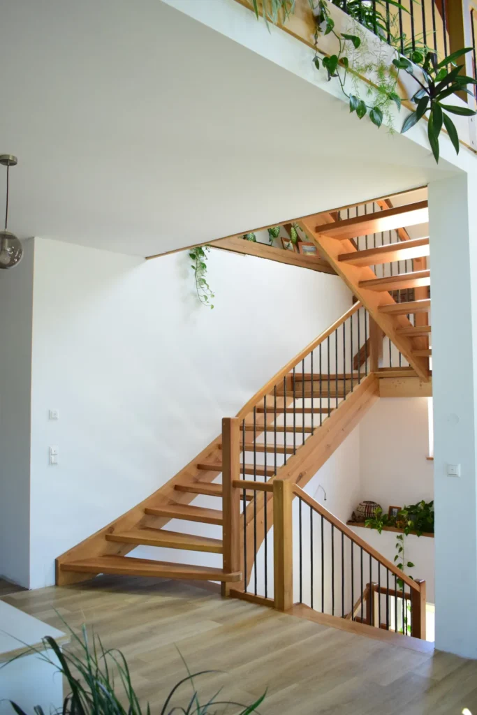 Eine Holztreppe mit schwarzem Metallgeländer in einem hellen, modernen Interieur. Grüne Pflanzen schmücken das Geländer und die Ecken, und auf der unteren Ebene ist ein heller Holzboden zu sehen. Natürliches Licht durchflutet den Raum.