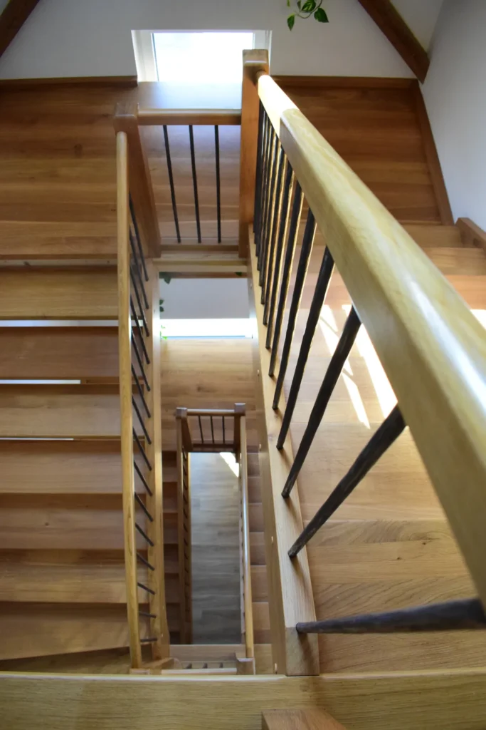 Eine Ansicht von oben auf eine Holztreppe mit schwarzen Metallgeländern, die durch das natürliche Licht aus einem kleinen Fenster ganz oben beleuchtet wird. Die Treppe scheint sich spiralförmig über mehrere Stockwerke hinabzuwinden.