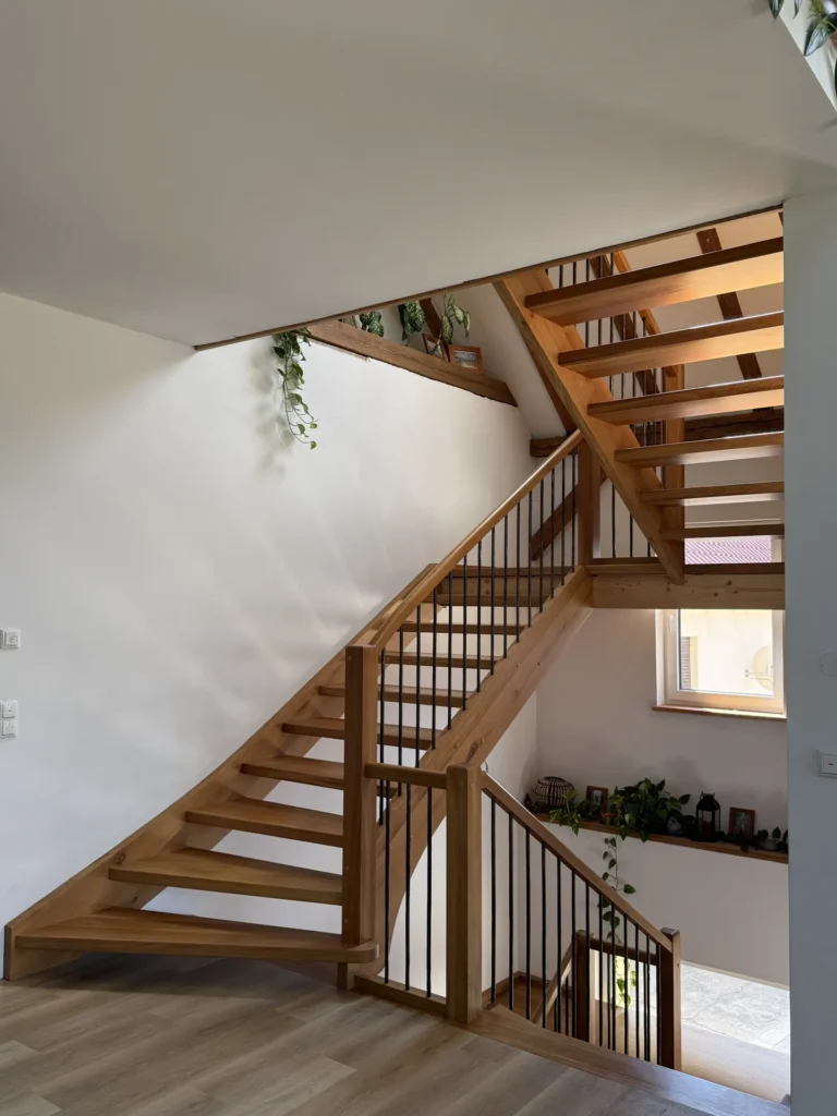 Eine moderne Holztreppe mit schwarzen Metallgeländern, Zimmerpflanzen auf den Fensterbänken und natürlichem Licht, das durch ein Fenster hereinströmt, vor dem Hintergrund weißer Wände und heller Holzböden.