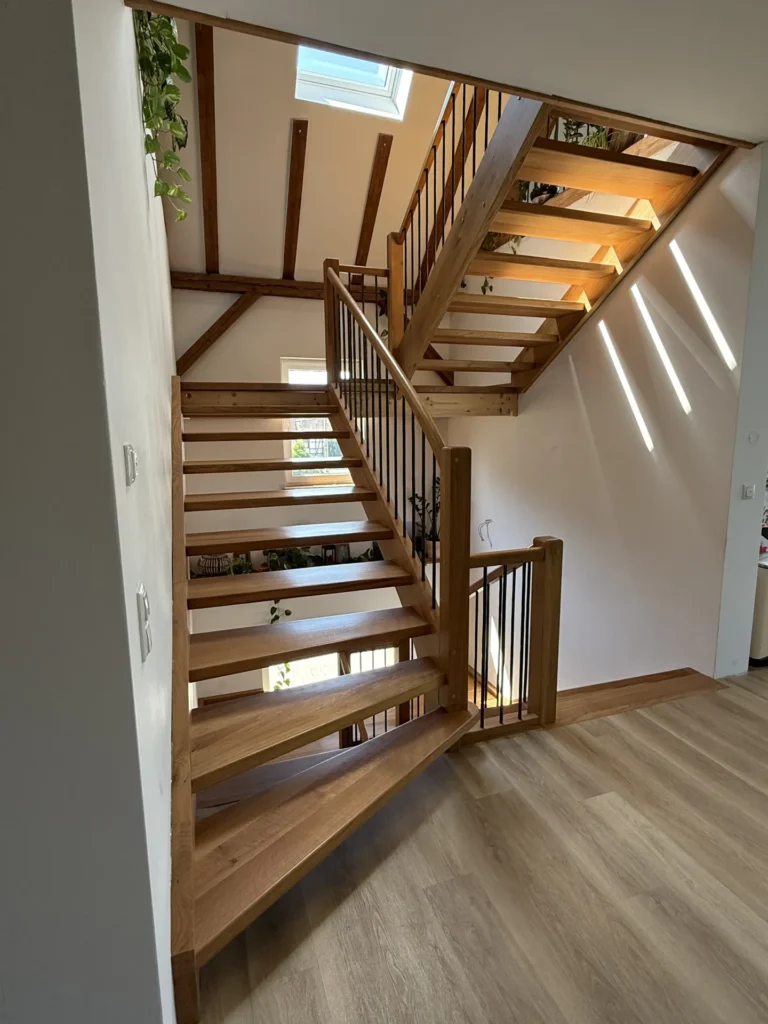 Eine Holztreppe mit offenen Setzstufen und Metallgeländern in einem modernen, hellen Interieur. Oberlichter und Sonnenstrahlen erhellen den Raum und lassen die Holzbalken und Pflanzen im Obergeschoss besonders gut zur Geltung kommen.