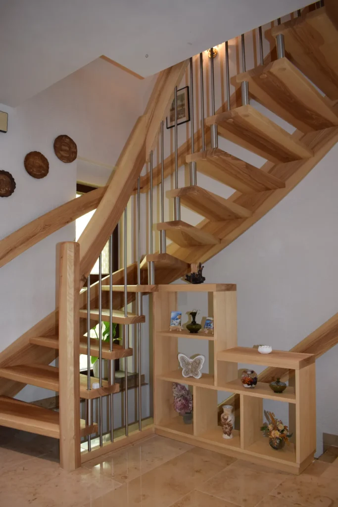 Eine Holztreppe mit Metallgeländer und offenen Setzstufen. Unter der Treppe befindet sich ein kleines Holzregal, auf dem Dekorationsgegenstände wie Vasen, Figuren und ein Kunstwerk in Form eines Schmetterlings ausgestellt sind. An den Wänden hängen einige runde Wandbehänge.
