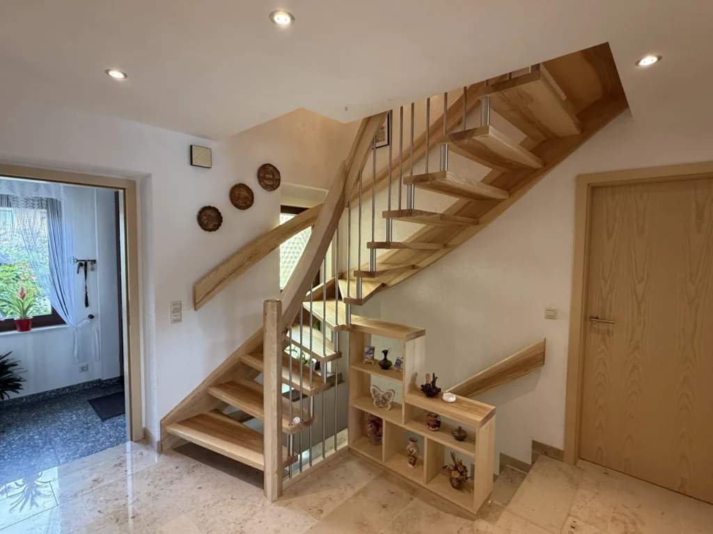 Holztreppe mit offenen Setzstufen und Metallgeländerstäben, unter der sich ein eingebautes Regal befindet, auf dem kleine Dekorationsgegenstände ausgestellt sind. Der Bereich ist durch Einbauleuchten an der Decke gut ausgeleuchtet. Links ist ein Flur mit Pflanzen zu sehen.
