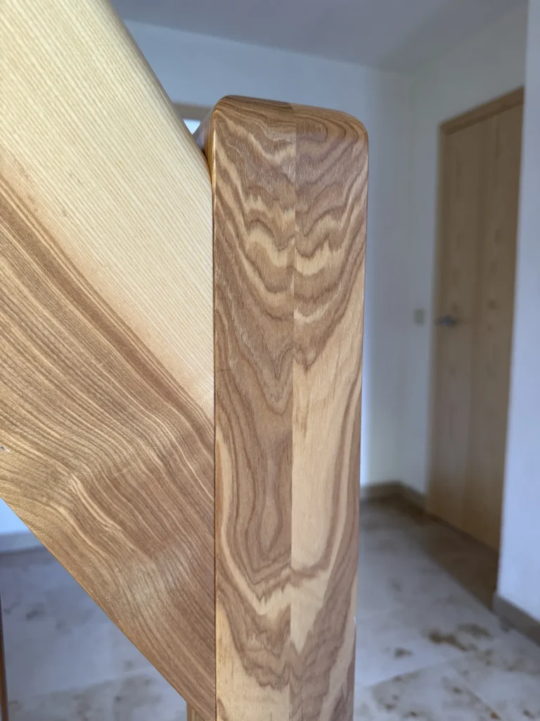 Nahaufnahme eines Holzgeländers mit sichtbarer Maserung in einem Raum mit Fliesenboden, weißen Wänden und einer geschlossenen Holztür im Hintergrund.