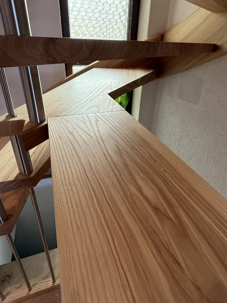 Nahaufnahme einer modernen Holztreppe mit sichtbarer Holzmaserung, Metallgeländern und einem Fenster mit Musterglas im Hintergrund.