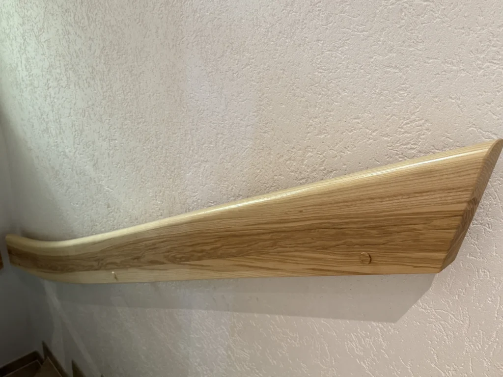 An einer strukturierten weißen Wand ist ein Handlauf aus poliertem Holz angebracht, der sich an einem Ende allmählich verjüngt.