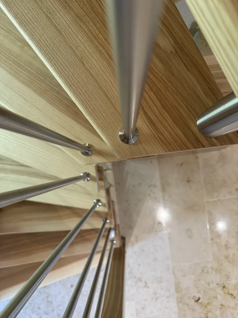 Nahaufnahme einer Holzwendeltreppe aus der Vogelperspektive mit Metallgeländern und einem Fliesenboden darunter. Das Bild hebt die geometrischen Linien und die Oberflächenstrukturen der Materialien hervor.