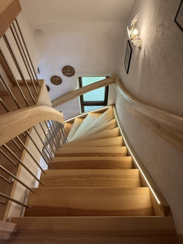 Holzwendeltreppe mit Metallgeländer, von oben gesehen. Durch ein Fenster am unteren Ende fällt natürliches Licht herein, und am Rand der Treppe sind warme Lichter angebracht. An der weißen, strukturierten Wand hängen runde Wanddekorationen.