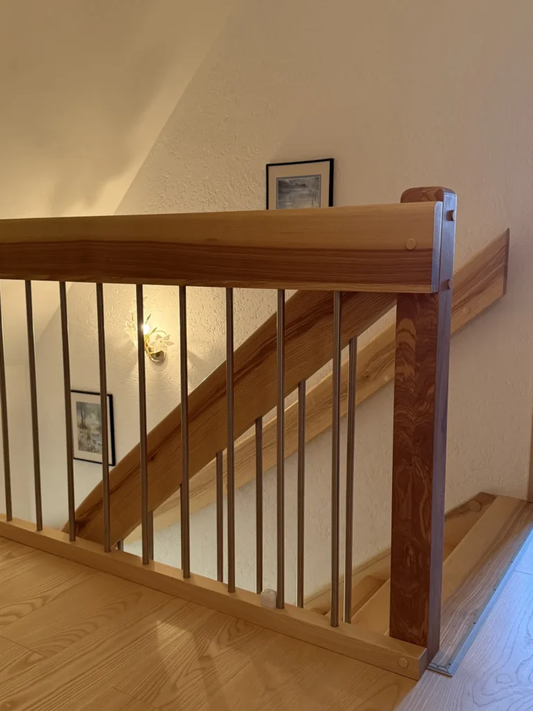 Eine Holztreppe mit vertikalen Metallbalustern und einem stabilen Handlauf. Ein heller Holzboden, gerahmte Bilder an der Wand und eine Wandlampe sorgen für eine gemütliche Atmosphäre im Raum.