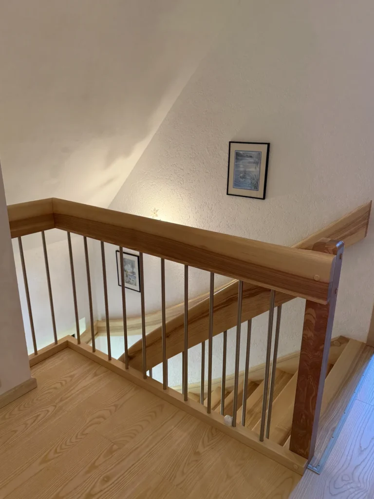 Eine Holztreppe mit Metallgeländer führt zwischen den Etagen hinab. An der weißen, strukturierten Wand hängen zwei gerahmte Bilder, und sanfte Beleuchtung erhellt den Raum. Das Foto wurde vom oberen Ende der Treppe aus aufgenommen.