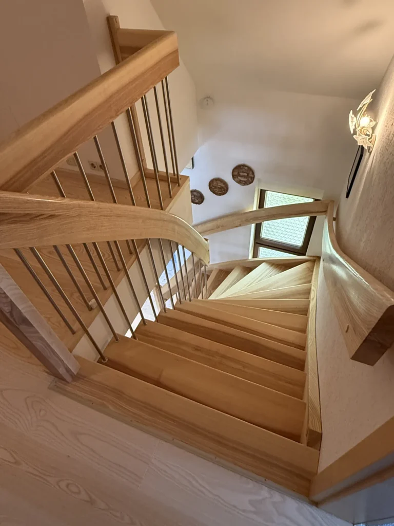 Eine Holztreppe mit Metallgeländer schlängelt sich in einem modernen Haus nach unten. Durch ein Fenster am Fuß der Treppe fällt natürliches Licht herein. Auf der rechten Seite sind dekorative Wandplatten und eine Wandleuchte zu sehen.