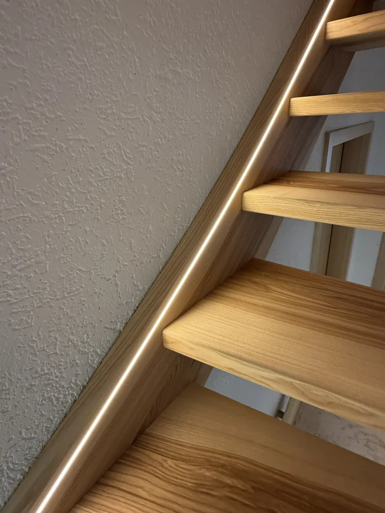 Holztreppe mit offenen Setzstufen und einem seitlich entlanglaufenden, eingebauten LED-Lichtband neben einer strukturierten weißen Wand.