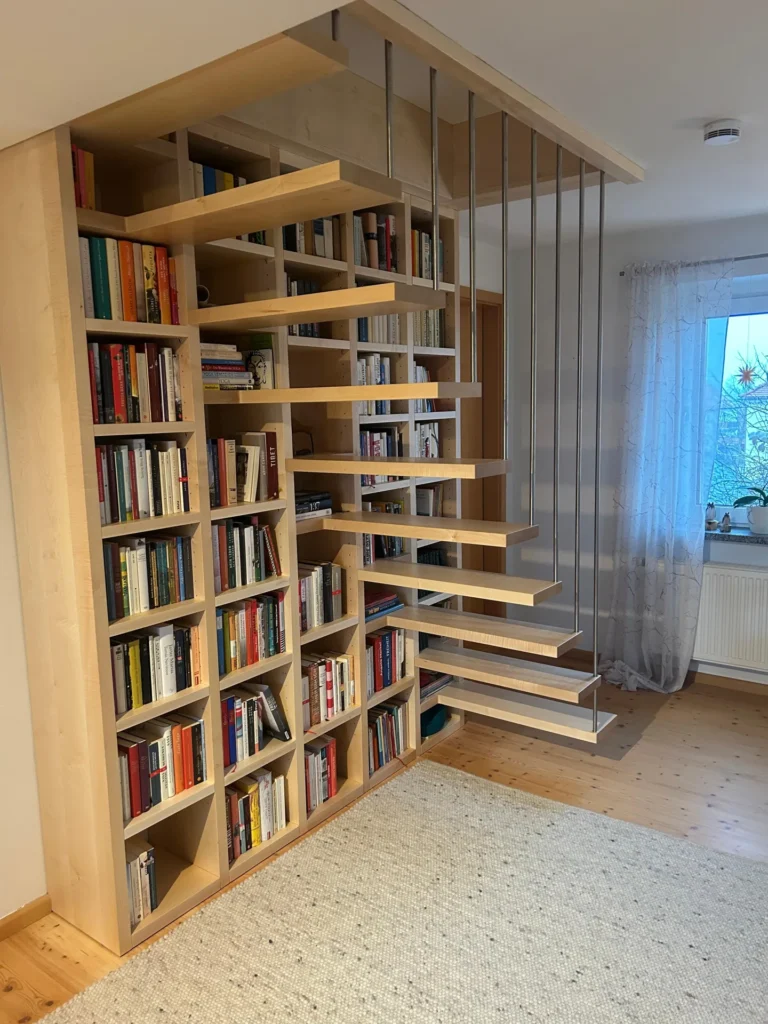 Eine moderne, schwebende Treppe mit offenen Holzstufen ist in ein Bücherregal voller Bücher eingebaut. Die Stufen werden von vertikalen Metallstangen gestützt und führen in das Obergeschoss. Natürliches Licht fällt durch ein Fenster mit transparenten Vorhängen herein.