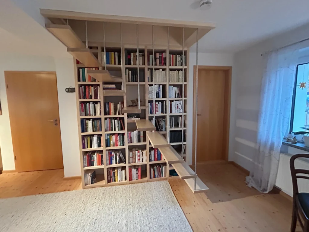 Eine Holztreppe mit offenen Stufen ist in ein raumhohes Bücherregal eingebaut, das mit Büchern gefüllt ist. Das Bücherregal unterteilt einen Raum mit Holzböden, Türen, einem Fenster mit weißen Vorhängen und einem cremefarbenen Teppich.