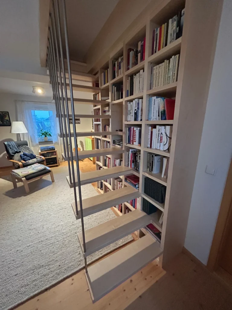 In einem gemütlichen, hell beleuchteten Wohnzimmer mit hellen Möbeln und einem hellen Teppichboden erstreckt sich neben einem hohen, mit Büchern gefüllten Bücherregal eine moderne Treppe mit offenen Holzstufen.