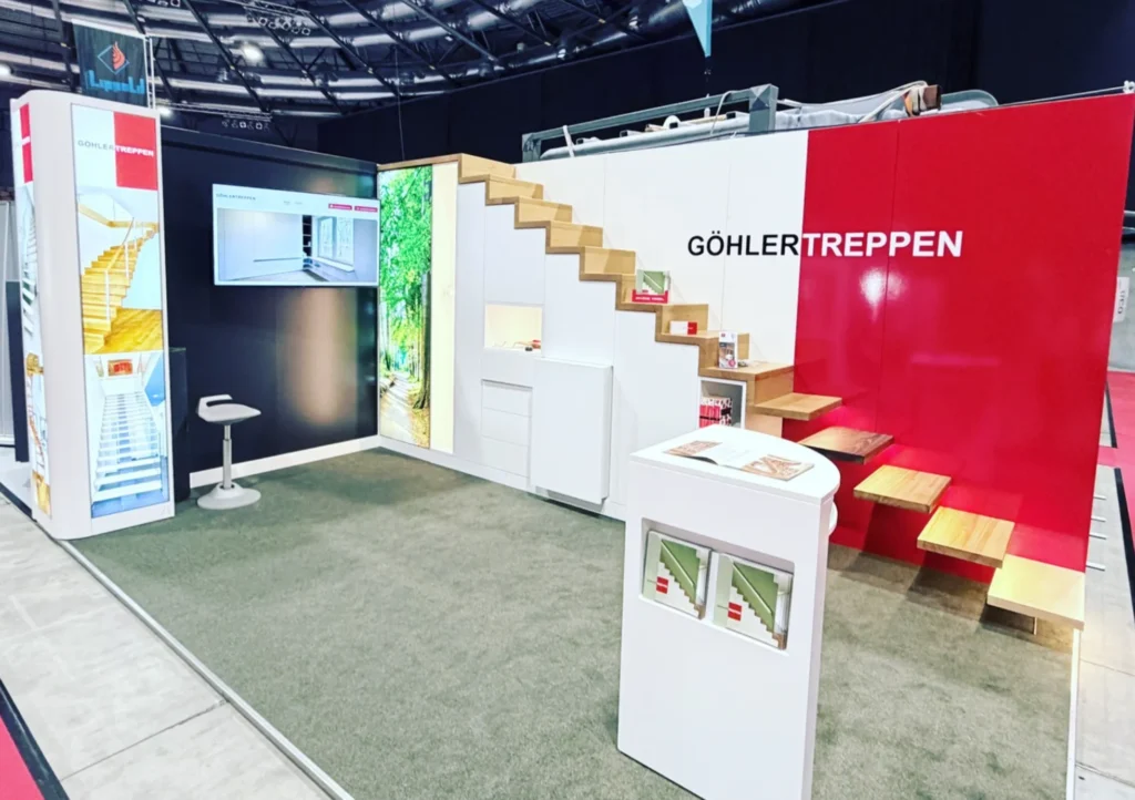 Ein moderner Messestand für GÖHLERTREPPEN präsentiert Holztreppen, weiße und rote Wände, einen Monitor, einen Prospektständer und eine kleine Sitzecke auf einem mit Teppichboden ausgelegten Boden.